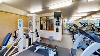 Located at 1040 NW 10th Ave in Portland's vibrant Pearl District, this fitness center is a well-equipped exercise room designed to support a complete workout experience within a compact, efficiently organized footprint. The space is outfitted with a diverse selection of cardio machines, including treadmills, ellipticals, and stationary bikes from brands such as True and Vectra, along with a multi-use strength training station and a workout bench. Large floor-to-ceiling mirrors line the walls, visually expanding the room and allowing users to monitor their form during exercise. Overhead fluorescent lighting ensures the space is bright and consistently illuminated throughout. Multiple windows along the exterior wall invite natural light and offer views of the surrounding Portland streetscape, adding an open, connected feel to the interior. The flooring is covered in durable, low-pile carpet with rubber mat sections beneath the equipment for stability and protection. A wall-mounted television provides entertainment during workouts, while a sanitizing station and posted informational signage support cleanliness and user guidance. Warm wood-toned trim accents the mirrors and doorframes, lending a polished finish to the otherwise functional design. The room is accessed through natural wood-finished doors, completing a clean and practical aesthetic throughout.