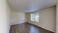 Located at 11721 E Nora Ave in Spokane Valley, WA, this two-bedroom, one-bathroom unit offers a thoughtfully arranged single-floor layout. Dark hardwood-style flooring flows throughout the main living areas and hallway, adding warmth and visual continuity. The living room is bright and inviting, featuring a large window that frames views of mature trees and greenery, complemented by a baseboard heating system and a textured ceiling. The kitchen is efficiently designed with warm wood cabinetry, laminate countertops, a double-basin sink, and a dishwasher, providing ample functionality for everyday use. The hallway connects the living spaces to the two bedrooms, each featuring neutral walls and dedicated closet space. The primary bedroom offers a generous footprint, while the second bedroom is finished with soft carpeting. The full bathroom includes a tub-shower combination with accessibility grab bars and hardwood-style flooring. A private patio extends the living space outdoors. The surrounding community grounds showcase expansive, well-manicured lawns, large shade trees, and a playground area, creating a serene and family-friendly environment.