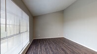 Located at 11511 Metric Blvd in Austin, TX, this single-floor unit offers a practical and well-organized layout designed for comfortable everyday living. Throughout the main living areas, rich dark hardwood-style flooring provides a warm, cohesive aesthetic that ties the space together.

The entry opens directly into a bright living area, where a large window fitted with horizontal blinds allows ample natural light to fill the room. Neutral gray walls and a textured ceiling complement the dark flooring, creating a clean and understated atmosphere. An additional room — suitable as a bedroom or secondary living space — mirrors this aesthetic with the same flooring, neutral tones, and a window for natural light.

The kitchen is accessed via an open pass-through layout and features dark cabinetry, a built-in dishwasher, black appliances including a range, and a breakfast bar-style counter that connects to the living area. Track lighting illuminates the kitchen corridor, and lighter wood-style flooring in this zone provides a subtle contrast to the rest of the unit.

The bathroom is clean and functional, featuring a full-size soaking tub with a tiled surround in crisp white, maintaining a simple and hygienic finish. A ceiling fan is also present, adding to the unit's overall comfort and ventilation.