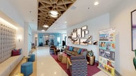 The leasing lounge at 986 Broadway in Chula Vista offers a polished and inviting environment designed to make a strong first impression. The single-floor space is anchored by an open main lounge area featuring a striking geometric wood-panel ceiling installation that adds warmth and architectural drama overhead. Recessed and pendant lighting complement the natural light that filters in through large floor-to-ceiling glass entry doors framed in sleek black steel, offering views of the exterior courtyard and lending the space an airy, open feel.

The lounge is furnished with contemporary seating arrangements, including a neutral-toned sofa accented with vibrant teal and pink throw pillows, stylish accent chairs, and a plush burgundy area rug that grounds the space with a pop of color. A gallery wall of beach-inspired photography in coordinating frames serves as a focal art feature, reinforcing a coastal aesthetic throughout. Polished tile flooring runs through the entry and main areas, transitioning to wood-toned flooring in quieter alcove seating zones.

A secondary seating nook features a chaise-style bench beneath a large framed print of a turquoise sea fan, adding a serene, nature-inspired accent. The overall ambiance is modern and energetic yet comfortable, with a cohesive coastal color palette of teal, white, and warm neutrals creating a welcoming atmosphere for prospective residents.