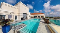 Located in the heart of Boca Raton, Florida, this rooftop pool deck at 401 Northeast Mizner Boulevard offers a refined outdoor retreat with a distinct Mediterranean architectural character. The space is defined by its cream-colored stucco walls, terra cotta-tiled rooflines, and decorative tilework accents that evoke a classic South Florida aesthetic.

At the centerpiece of the deck sits a generously sized swimming pool with crystal-clear turquoise water, complemented by a separate spa for relaxation. The surrounding deck is finished in light-toned tile, creating a clean, resort-like atmosphere that reflects the abundant Florida sunshine. A decorative arched niche with intricate mosaic tilework serves as a striking architectural focal point along the pool's edge.

Lounge seating and sun chairs are arranged throughout the deck, inviting residents to take in the open sky and sweeping rooftop views of the surrounding Boca Raton landscape. A bar or service area is also present, enhancing the space's amenity-rich character. The elevated position of the deck allows for unobstructed views under broad blue skies, further amplifying the sense of openness and leisure. This rooftop amenity space combines elegant design with functional outdoor living in one of South Florida's most desirable addresses.
