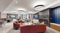 This generously sized communal living space offers a thoughtfully designed, open-plan layout that seamlessly blends multiple lounge and social zones. The interior showcases a contemporary aesthetic defined by light-toned luxury vinyl plank flooring, crisp white ceilings, and bold accent walls in deep navy blue with decorative paneling detail. Signature circular pendant light fixtures are suspended throughout, providing warm, layered illumination that complements the recessed ceiling lighting.

At the heart of the space, a sophisticated seating area features a curved terracotta sofa, modular grey sectionals, and cream accent chairs arranged around round coffee tables. A dramatic floor-to-ceiling grey marble fireplace serves as a striking focal point, flanked by a built-in media wall with a large flat-screen TV and vertical wood slat paneling. Adjacent to the main lounge, multiple casual dining clusters with round tables and upholstered chairs provide flexible spaces for gathering or working.

Warm wood-clad columns anchor the layout and add natural texture throughout. Large windows allow natural light to filter into the space, enhancing its open, airy atmosphere. A transitional corridor area with wood-paneled ceilings and a staircase with glass railings connects to adjacent amenity spaces, completing this well-appointed, multi-functional communal environment.