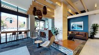 This single-floor space is designed as a sophisticated communal lounge, blending modern design elements with warm, inviting touches. The layout is organized into distinct seating zones, each offering a different atmosphere while maintaining a cohesive aesthetic throughout.

At the heart of the space sits a striking tiled fireplace, clad in a mosaic of blue, gray, and brown tiles, serving as a natural focal point. Surrounding it, contemporary sculptural chairs and a small accent table create an intimate conversation area. Overhead, a series of layered natural wood slat ceiling panels add architectural interest and warmth, complemented by dramatic woven pendant lights in an organic teardrop form.

Natural light filters in through large windows along one wall, where a counter-height bar seating area provides a casual workspace. A deeper lounge zone to the right features plush booth-style seating set against a rich teal accent wall adorned with framed artwork.

The flooring transitions between tile and patterned carpet, subtly defining each zone. Lush greenery and carefully chosen décor contribute to a coastal-inspired, relaxed ambiance, making the space both functional and visually dynamic.