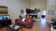 This single-floor space is defined by its striking high ceilings, which lend an airy, expansive feel to the open-concept layout. Crown molding runs along the top of the walls, adding a refined architectural detail that complements the neutral, warm-toned walls throughout.

The living area is anchored by a rich burgundy sofa adorned with decorative throw pillows, paired with a glass-top coffee table. Warm hardwood flooring extends throughout the space, providing visual continuity between the living, dining, and kitchen zones.

The kitchen features dark espresso cabinetry extending to near ceiling height, topped with decorative greenery. A dark granite or stone countertop provides workspace, and stainless steel appliances — including a refrigerator and microwave — offer modern functionality. A water cooler is positioned nearby, adding a practical touch.

Adjacent to the kitchen, a compact dining area accommodates a small table with patterned upholstered chairs, serving as a natural transition between cooking and living spaces.

Decorative wall art and potted plants are thoughtfully placed throughout, contributing to a cohesive and welcoming ambiance. The overall design balances functionality with comfort in a well-organized, open layout.