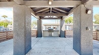 Located at 8560 W Peoria Ave in Peoria, Arizona, this single-level outdoor ramada serves as a communal gathering space designed for open-air relaxation and entertaining. The structure is defined by its striking vaulted, exposed wood-beam ceiling, which provides generous shade while maintaining an airy, open feel on all sides. Sturdy textured concrete columns anchor the pavilion at its corners, supporting the pitched roof and framing views of the surrounding residential community and mature trees beyond.

The concrete slab floor extends throughout the covered area, offering ample space for outdoor furniture and social gatherings. A standout feature of the ramada is the centrally positioned built-in stainless steel BBQ grill station, providing a dedicated outdoor cooking area ideal for community events and cookouts. Horizontal wood slat fencing lines the perimeter, adding a warm, contemporary contrast to the rugged concrete elements and helping to define the space without fully enclosing it.

Natural light floods the area from all open sides, creating a bright and inviting atmosphere. The ramada's open-air design takes full advantage of the Arizona climate, while the covered roof ensures shade and comfort during sunny days. Concrete waste receptacles are thoughtfully placed throughout the space. The overall design balances functional utility with a casual, community-focused aesthetic, making it a practical and welcoming outdoor amenity.