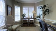 This single-story space showcases a well-appointed sitting or study area defined by refined architectural elements and a warm, inviting ambiance. Crown molding and chair rail detailing along the walls add a classic, polished character to the interior. Three large windows dressed with white plantation shutters allow generous natural light to filter into the room while maintaining privacy, and glimpses of lush greenery outside hint at a pleasant outdoor setting. The neutral taupe wall color provides a versatile backdrop that complements the room's mix of traditional and industrial-inspired furnishings. A rustic wood-and-metal shelving console serves as a functional centerpiece, styled with woven basket storage, decorative dishes, and a tall table lamp. Flanking the console are potted tropical plants that introduce organic texture and life to the space. Seating includes a classic upholstered arm chair with nailhead trim and an additional chair partially visible at the edge of the frame, suggesting a comfortable, functional layout. Neutral carpeting grounds the room, contributing to its overall soft and cohesive aesthetic.