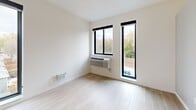 Located on Minnehaha Ave in Minneapolis, Minnesota, this single-floor unit presents a clean, contemporary design characterized by a crisp all-white palette and light wood-tone flooring that runs consistently throughout the space. The open layout, visible in the overhead model view, reveals a well-organized arrangement that includes a main living area, a kitchen, and a bathroom.

The living and sleeping areas feature smooth white walls and light hardwood-style flooring, creating a bright and airy atmosphere. Tall, narrow black-framed windows allow natural light to filter in while offering views of the surrounding neighborhood greenery. A wall-mounted HVAC unit provides climate control, and electrical panels are thoughtfully integrated into the walls.

The kitchen is outfitted with modern cabinetry and built-in appliances, maintaining the unit's minimalist aesthetic. A hallway lined with floor-to-ceiling white cabinetry provides generous storage with sleek chrome hardware, leading toward the bathroom.

The bathroom is finished with light wood-tone flooring, a dark-toned vanity with a white countertop and integrated sink, a wall-mounted mirror, and a full tub-shower combination with a chrome curtain rod. A frosted-glass door adds a refined, modern touch. Overall, the space reflects a cohesive, contemporary design sensibility with functional, well-appointed details throughout.