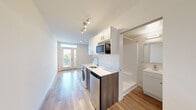 Located at 3300 NE 125th St in Seattle's Pinehurst neighborhood, this efficiently designed studio offers a thoughtfully organized single-floor layout that maximizes every square foot. Light wood-look flooring runs throughout the space, complementing crisp white walls and a clean, contemporary aesthetic.

The open main area flows directly from the entrance, where a glass-paneled door with a transom window and sidelight fills the room with natural light and offers views of the surrounding greenery. The compact kitchenette is smartly outfitted with white upper cabinetry, a microwave, a single-basin sink with a modern faucet, a stainless steel mini-refrigerator, and an in-unit washer — providing essential conveniences within a minimal footprint. A built-in closet rod offers dedicated storage space adjacent to the kitchen area.

The bathroom features a clean, modern design with a wall-mounted vanity, a large mirror, a walk-in shower, and wood-look flooring consistent with the rest of the unit. A sliding door separates the bathroom from the main living space, contributing to an open and airy feel. Track lighting in the kitchen area and ample natural light from the entrance door ensure the space remains bright and welcoming throughout.