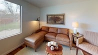 This single-floor space showcases a cohesive, nature-inspired design carried consistently across each room through warm wood flooring, earthy tones, and thoughtfully chosen decor. The living area serves as the heart of the space, centered around a rich cognac leather sectional sofa paired with a striped accent chair, both arranged on a woven area rug. A live-edge wood coffee table anchors the seating arrangement, adorned with decorative sculptural accents. Framed botanical and wood-themed artwork adds visual warmth to the soft, neutral walls, while a floor lamp and table lamp provide layered ambient lighting. Natural light filters in through a window, enhancing the inviting atmosphere. The compact kitchen is efficiently laid out, featuring granite-toned countertops, white cabinetry with gold trim accents, and white appliances including a full-size refrigerator and electric range with overhead ventilation. The hardwood flooring continues seamlessly into the bathroom, which includes a granite-topped vanity, a bathtub with a striped fabric shower curtain in warm reds and golds, and decorative wall art crafted from natural wood slices. Overall, the space presents a warm, cohesive aesthetic with rustic natural elements throughout.