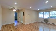 This single-story space showcases a clean, functional design with well-maintained hardwood floors that extend throughout the main living area, lending warmth and a classic aesthetic to the interior. The open layout provides a generous sense of space, allowing for flexible furniture arrangements to suit a variety of needs.

Recessed lighting is installed throughout the ceiling, offering consistent, even illumination across the room, while a ceiling fan adds both a practical and decorative element. The neutral, light-toned walls create a versatile backdrop that complements a range of décor styles.

Natural light enters through multiple windows, including one that frames an outdoor view of surrounding greenery, contributing to the bright and airy feel of the space. Baseboard heaters are positioned along the walls, providing efficient heating throughout the colder months.

A doorway leads to an adjacent room, visible in the background, which features its own window and suggests additional usable space within the floor plan. The white trim and clean baseboards throughout add a crisp, finished look, rounding out the overall well-kept and move-in-ready presentation of this single-floor property.