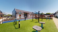 Located at 7913 Harwood Rd in North Richland Hills, TX, this outdoor amenity space is a well-equipped fitness and recreation area designed to promote an active lifestyle. Set against the backdrop of a multi-story brick residential building with distinctive teal accent doors and warm wood panel detailing, the space combines functionality with an inviting community atmosphere.

The grounds are covered in lush artificial turf, providing a clean, low-maintenance surface suitable for year-round use. A dedicated sprint and agility track is prominently featured, marked with lane lines and the motivational phrase "Live Life. Move Strong." — underscoring the space's commitment to wellness.

The outdoor gym area includes a variety of equipment such as a pull-up and suspension training rig, resistance stations, heavy tire sleds, and freestanding rotational fitness stations, offering a comprehensive full-body workout experience without the need for an indoor facility.

Communal seating is thoughtfully incorporated, with wooden benches positioned along a raised platform area, encouraging social interaction. Concrete walkways border the space, while neatly trimmed shrubs and gravel landscaping along the building's exterior add a polished, well-maintained appearance. Overhead shade structures further enhance comfort during sunny Texas days, making this a highly functional and appealing outdoor fitness destination.