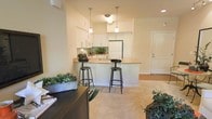 This single-floor residence offers a thoughtfully designed open-concept layout that promotes a sense of spaciousness and ease of movement. The kitchen is a standout feature, outfitted with crisp white cabinetry, light granite countertops, and full-size appliances. A peninsula breakfast bar with seating for two serves as both a functional workspace and a casual dining area, anchored by a pair of stylish pendant lights that add a warm, inviting glow. The kitchen flows seamlessly into the living area, which is finished with soft, neutral-toned carpeting that lends a comfortable and welcoming ambiance. Walls painted in a warm off-white tone unify the space and reflect the abundant recessed and natural lighting. Greenery and potted plants are thoughtfully placed throughout, adding life and a touch of nature to the interior. The entryway, visible from the main living area, features a solid front door with clean architectural trim, providing a polished transition between the interior and exterior. The overall design balances practicality with aesthetic appeal, making the space feel both functional and inviting for everyday living.