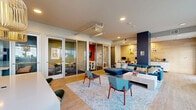 Located at 2050 Durant Ave in Berkeley, California, this contemporary communal lounge offers a well-appointed, single-floor space designed for both relaxation and collaboration. The open floor plan is anchored by light hardwood flooring that runs throughout, contributing to a warm and cohesive aesthetic.

The main living area is furnished with a navy blue sectional sofa, colorful accent pillows, and mid-century modern style chairs in coordinating blue tones, all arranged around a sleek white coffee table atop a geometric patterned area rug. Bold navy accent columns punctuate the space, adding architectural interest and visual contrast against the neutral walls.

A dedicated work and dining nook features a white communal table topped with pendant lights crafted from natural woven materials, paired with sage green chairs — a stylish spot suited for casual meetings or focused work. Adjacent glass-panel sliding doors lead to enclosed private rooms, ideal for smaller group gatherings or quieter activities, and are equipped with their own seating and wall-mounted displays.

Large windows along multiple walls flood the space with natural light and offer views of the surrounding Berkeley neighborhood. Colorful curated artwork is displayed throughout, enhancing the vibrant, creative atmosphere. Recessed lighting and exposed duct details on the ceiling add an industrial-modern touch, rounding out the thoughtfully designed environment.