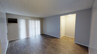 This single-floor space offers a clean, modern aesthetic defined by its neutral tones and practical design. The main living area is an open, airy room finished with rich, dark hardwood-style flooring that runs throughout, creating a warm contrast against the soft gray walls and bright white trim. A large sliding glass door fitted with vertical blinds dominates one wall, allowing generous natural light to flood the space and lending a sense of openness. The textured ceiling adds a subtle architectural detail overhead. Adjacent to the main living area, a well-lit entryway or hallway is visible, finished with lighter-toned flooring that provides a clean transition between spaces. The corridor leads to additional rooms beyond, suggesting a functional and straightforward layout. The overall ambiance of the space is fresh and move-in ready, with a minimalist character that provides a versatile canvas for a variety of interior styles. The combination of natural light, neutral finishes, and durable flooring makes this a practical and comfortable single-level space.