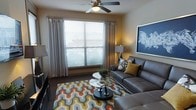 This single-floor living space offers a thoughtfully designed interior centered around a comfortable and visually engaging living area. The room is anchored by a large window dressed with horizontal blinds and flowing drape panels, allowing generous natural light to fill the space while maintaining privacy options. A ceiling fan with an integrated light fixture is positioned centrally overhead, contributing both functional air circulation and a subtle design element.

The living area is furnished with a tufted gray leather sofa set that provides a sleek, contemporary feel. A round glass-top coffee table with a dark pedestal base serves as the focal point of the seating arrangement, complemented by a bold geometric-patterned area rug in shades of orange, gold, gray, and white that adds visual warmth and energy to the space. Decorative throw pillows in coordinating tones enhance the cohesive color palette.

Wall décor includes a framed blue-toned artwork depicting an expressive figure, adding an artistic accent to the neutral warm-toned walls. A table lamp in the corner provides ambient lighting. The overall ambiance is modern, inviting, and well-coordinated, balancing comfort with contemporary style.