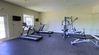 This fitness center offers a practical and functional workout environment designed to support a range of exercise routines. The space is laid out on a single floor with durable rubber flooring that provides excellent traction and cushioning underfoot — a key feature for any exercise facility. The walls are finished in a clean, neutral tone, contributing to a bright and open atmosphere.

The cardio section is well-represented, featuring multiple treadmills that allow for simultaneous use, making the space suitable for shared or community use. A rowing machine rounds out the cardio options, offering a full-body, low-impact workout alternative. Natural light filters in through a window along one wall, enhancing the overall ambiance and creating a more inviting exercise environment. A wall-mounted flat-screen television is also present, providing entertainment during workouts.

On the strength training side, a multi-station cable and weight machine dominates the right side of the room, offering a versatile range of exercises targeting multiple muscle groups from a single unit. The equipment is well-organized, leaving sufficient open floor space for movement between stations. Overall, the fitness center is a streamlined, efficiently arranged facility that provides the essentials for both cardiovascular and strength-based training.