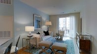 This single-floor residence offers a thoughtfully designed living space that blends comfort with contemporary elegance. The living room serves as the centerpiece of the home, bathed in natural light that streams through large sliding glass doors leading to a private balcony, offering views of the surrounding urban streetscape. Sheer floor-to-ceiling curtains frame the doors gracefully, enhancing the airy, open ambiance of the room. The walls are finished in a soft, calming blue-gray tone that complements the bright white upholstered sofa and coordinating accent pieces. A patterned blue area rug anchors the seating arrangement, which includes a classic armchair and a pair of tufted ottomans that add both style and functionality. A glass-top console table along one wall holds elegant lamp and decorative accents, while a framed wall art piece adds a curated, gallery-like quality to the space. Decorative table lamps provide warm ambient lighting to complement the natural light. The overall aesthetic is clean, refined, and welcoming, with a coastal-inspired color palette that creates a serene and livable atmosphere throughout.