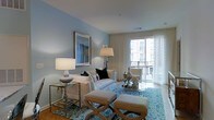 This single-floor residence offers a thoughtfully designed living space that blends comfort with contemporary elegance. The living room serves as the centerpiece of the home, bathed in natural light that streams through large sliding glass doors leading to a private balcony, offering views of the surrounding urban streetscape. Sheer floor-to-ceiling curtains frame the doors gracefully, enhancing the airy, open ambiance of the room. The walls are finished in a soft, calming blue-gray tone that complements the bright white upholstered sofa and coordinating accent pieces. A patterned blue area rug anchors the seating arrangement, which includes a classic armchair and a pair of tufted ottomans that add both style and functionality. A glass-top console table along one wall holds elegant lamp and decorative accents, while a framed wall art piece adds a curated, gallery-like quality to the space. Decorative table lamps provide warm ambient lighting to complement the natural light. The overall aesthetic is clean, refined, and welcoming, with a coastal-inspired color palette that creates a serene and livable atmosphere throughout.