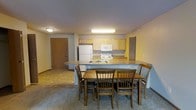This single-floor space offers a practical and functional layout centered around an open-concept kitchen and dining area. The kitchen is equipped with light wood cabinetry, a white refrigerator, electric range, microwave, and a double-basin sink set within a wraparound countertop that doubles as a breakfast bar — creating a seamless transition between cooking and dining. The countertops feature a neutral laminate finish that complements the warm tones of the cabinetry. Adjacent to the kitchen, the dining area accommodates a wooden table with seating for four, making it well-suited for shared meals or casual gatherings. Overhead fluorescent lighting in the kitchen provides ample task illumination, while flush-mount fixtures extend consistent lighting throughout the space. The flooring transitions from tile near the entryway to carpet in the main living areas, adding a layer of texture and comfort underfoot. A solid wood entry door opens directly into the common area, offering a straightforward and accessible floor plan. The overall ambiance is warm and utilitarian, with neutral wall tones and a no-frills design that prioritizes ease of use and livability.