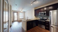 This single-floor residence is designed with a modern aesthetic and functional layout. The open-concept kitchen serves as a central feature, showcasing rich dark cabinetry extending to the ceiling, granite countertops, and a full suite of stainless steel appliances including a range, microwave, and refrigerator. A mosaic tile backsplash adds visual interest, while a peninsula with a granite surface and built-in sink provides additional workspace and separates the kitchen from the living area. Track lighting and hardwood flooring carry throughout the main living space, which benefits from natural light streaming through windows and a glass door leading to a balcony. A ceiling fan keeps the living area comfortable. A hallway connects to the remaining rooms, including a carpeted bedroom featuring dual windows with city views and its own ceiling fan. The bathroom is well-appointed with dark cabinetry, a granite-topped vanity, a framed mirror with overhead lighting, and tile flooring. A dedicated laundry area with washer and dryer hookups adds convenience. The overall ambiance is contemporary and cohesive, with consistent finishes throughout.