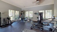 Located at 9989 Dorchester Rd in Summerville, South Carolina, this fitness center is a well-appointed, single-floor facility designed to support a comprehensive workout experience. The space is finished with durable rubber flooring and soft gray walls accented by crisp crown molding, creating a clean and motivating environment throughout.

Natural light pours in generously through multiple large windows that frame views of the surrounding outdoor area, including a glimpsed pool and lush greenery. A skylight adds additional brightness to the central area, while ceiling fans ensure comfortable air circulation during workouts.

The cardio zone is equipped with multiple treadmills featuring integrated display screens, an elliptical trainer, and a stationary bike, all positioned to take advantage of the natural light and outdoor views. A large floor-to-ceiling wall mirror amplifies the sense of space and allows for form monitoring during exercise.

The strength training area offers a diverse selection of equipment, including a multi-station cable machine, a VR1 strength machine, adjustable benches, and free weights including kettlebells and dumbbells. The variety of machines supports both upper and lower body training routines.

Overall, the fitness center presents a bright, spacious, and fully equipped environment suitable for a wide range of fitness activities, making it a functional and inviting amenity for its users.