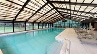 This indoor aquatic facility is housed within a standout glass-and-steel enclosure designed to flood the interior with natural light while providing a year-round swimming environment. The structure's pitched, translucent roof panels span the full length of the space, supported by a bold dark steel framework that gives the facility a clean, architectural character.

The centerpiece is a generously sized swimming pool with clear turquoise water, accessible via stainless steel railings and steps. Depth markers are visible along the pool edge, and the wide surrounding deck provides ample circulation space for swimmers and guests alike.

A dedicated relaxation zone runs alongside the pool, furnished with multiple sling-style lounge chairs and small side tables — ideal for unwinding before or after a swim. Additional seating arrangements, including chairs and bistro-style tables, are positioned near the glass perimeter walls, allowing guests to enjoy views of the lush green lawn and mature trees outside.

Brick accent walls add warmth and texture to the interior, contrasting with the predominantly glass enclosure. Floor-to-ceiling windows and sliding glass doors run along multiple sides of the structure, connecting the interior to the outdoors and ensuring the space feels open and airy. An exterior staircase provides direct access to the surrounding grounds, making this facility both functional and inviting.