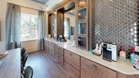Located at 18 Linestowe Drive in Belmont, NC, this thoughtfully designed single-floor space combines sophisticated aesthetics with functional versatility. The layout, as revealed in the floor plan, encompasses distinct zones that transition seamlessly from one to the next, creating a cohesive and inviting environment throughout.

The reception or front office area immediately impresses with soaring vaulted ceilings finished with warm wood paneling and a striking gold chandelier. A large grid-pane interior window wall connects to an adjacent private office, while curated heron artwork and sconce lighting contribute a refined, gallery-like atmosphere. A rustic wood desk anchors the space with understated elegance.

A dedicated service or bar area features an expansive run of rich walnut cabinetry with glass-front uppers, white quartz countertops, decorative honeycomb tile backsplash, and hardwood flooring — offering both practicality and visual depth. A coffee station adds a welcoming, hospitality-forward touch.

The main lounge area is generously proportioned, with dark hardwood floors, layered area rugs, and curated seating including a velvet chaise and tufted wingback chairs. Multiple windows dressed in linen drapes provide ample natural light and outdoor views. A wall-mounted television, burl wood coffee table, and carefully selected artwork complete this warm, eclectic interior.