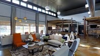 This single-floor space showcases a thoughtfully designed open-concept layout that seamlessly blends a living area with a kitchen and bar space. The architecture is defined by dramatically high ceilings finished in deep charcoal, creating a striking contrast with the bright, airy atmosphere generated by an expansive band of clerestory windows that run along the upper perimeter of the space. These elevated windows flood the interior with abundant natural light while maintaining privacy at eye level.

The living area is anchored by a neutral-toned sofa adorned with layered throw pillows in hound's-tooth and solid patterns, complemented by bold orange accent chairs that inject vibrant energy into the palette. A rustic wood coffee table grounds the seating arrangement atop a textured area rug.

At the rear, a pergola-style architectural element frames the kitchen and bar area, adding a distinctive indoor-outdoor aesthetic. Dark cabinetry, pendant lighting, and bar seating define this functional zone. Large roller shades on the surrounding windows offer flexible light control. The overall ambiance is contemporary yet warm, blending industrial structure with inviting furnishings.