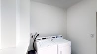 This single-floor space includes a functional laundry area designed to accommodate full-sized laundry appliances. The laundry nook houses a side-by-side top-loading washer and front-loading dryer, both in white, providing a complete in-unit laundry solution. The walls are finished in a light, neutral tone that contributes to a clean and utilitarian atmosphere. Utility connections, including water hoses and an electrical outlet, are visibly accessible along the back wall, indicating proper infrastructure for laundry use. The space is compact yet efficiently arranged to make the most of the available area, with smooth walls and a simple layout that keeps the focus on functionality. The minimalist design of this laundry area ensures ease of use and straightforward maintenance.