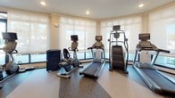 This modern fitness center is thoughtfully designed to accommodate a full range of workout styles across a single, open floor. The space is anchored by large windows along multiple walls, flooding the interior with natural light and offering views of the surrounding outdoor environment. Roller shades provide flexibility for light control throughout the day.

The cardio zone features a strong lineup of equipment including treadmills, stationary bikes, and stair climbers, each equipped with individual display screens. Adjacent to this area, a dedicated strength training section houses a variety of resistance machines, cable systems, a functional trainer, and a well-stocked dumbbell rack with an accompanying adjustable bench.

A standout feature of the fitness center is the functional turf training area, outfitted with balance tools such as a stability ball and BOSU trainer, ideal for dynamic and sport-specific workouts. Mounted flat-screen televisions are positioned throughout the space for entertainment during exercise.

The facility includes a water station for hydration convenience, recessed ceiling lighting, and a clean, neutral color palette with white walls and speckled rubber flooring that contribute to a professional and energizing atmosphere. Entry is accessible through both a glass-panel side door and a set of double doors, ensuring easy access from multiple points.