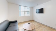 Located at 815 S Aurora St in Ithaca, NY, this three-bedroom, one-bathroom unit offers a thoughtfully designed single-floor layout with a clean, modern aesthetic. The open-concept kitchen and living area serves as the heart of the unit, featuring light wood-style flooring, white shaker cabinetry, stainless steel appliances including a refrigerator, range, and microwave, and a striking yellow backsplash that adds a pop of color. A peninsula with bar seating and a pendant light above enhances functionality and style. The living area accommodates comfortable seating, a wall-mounted TV, and a round dining table, all bathed in natural light from large windows fitted with blinds. A central hallway connects the three bedrooms, each carpeted in a neutral tile-pattern carpet and equipped with individual windows that bring in natural light. The bedrooms are compact yet functional, offering space for a bed and desk setup. The bathroom features wood-style flooring and a clean, minimal design with white walls. Throughout the unit, white panel doors and crisp white walls maintain a bright, cohesive feel, making this a practical and contemporary living space.