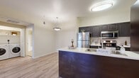 Located at 9971 E Speedway Blvd in Tucson, Arizona, this single-floor residence offers a well-designed, functional layout with a clean, contemporary aesthetic throughout. The kitchen serves as a central highlight, featuring dark espresso-toned cabinetry with arched door detailing, a suite of stainless steel appliances including a refrigerator, microwave, and range, and a peninsula with a white countertop and undermount sink — creating a natural divide between the kitchen and the adjoining living area. Pendant and flush-mount lighting illuminate the space effectively. Light wood-style flooring runs through the main living areas and bedrooms, lending a warm, cohesive feel across the unit. The living room is bright and airy, benefiting from natural light through large windows and a glass door, and is equipped with a ceiling fan. The bedrooms are generously proportioned with neutral wall tones and ample space. The bathroom is finished with tile flooring, a full tub-shower combination, and a vanity with a light countertop. A dedicated in-unit laundry area accommodates a full-size, front-loading washer and dryer set with overhead shelving for additional storage. The overall design is practical and well-appointed, suited for comfortable everyday living.