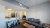 This single-floor unit offers a functional and well-organized layout across several distinct areas. The open-concept main living space combines the living room, dining area, and kitchen in one connected zone. The living room is furnished with leather seating, a coffee table, and a flat-screen TV, with a window providing natural light and outward views. A dining table with chairs sits nearby, creating a seamless transition between the two areas.

The kitchen is equipped with light maple cabinetry, granite-style countertops, and black appliances including a refrigerator, range, microwave, and dishwasher. A peninsula with bar seating adds additional counter space and separates the kitchen from the living area. Wood-look flooring runs throughout the kitchen and dining spaces.

Two carpeted bedrooms each feature windows with blinds for natural light and privacy. The bathroom includes a single-sink vanity with granite-style countertops, a shower with glass enclosure, and tile flooring. An attached walk-in closet provides built-in shelving and drawer storage. A dedicated laundry room houses a full-size washer and dryer with wall-mounted shelving above.