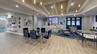 Located at 4300 Horizon North Pkwy in Dallas, TX, this single-floor community clubhouse offers a sophisticated and welcoming environment designed for relaxation and social interaction. The space is unified by a bold, blue-and-silver patterned carpet that anchors the various zones, complemented by light wood-style flooring in key areas.

The main lounge area features a pair of plush gray sofas arranged around a mirrored glass coffee table, with a large wall-mounted flat-screen TV set against an illuminated wave-textured accent wall with built-in greenery — creating a striking focal point. Ambient teal LED lighting adds a vibrant, modern touch throughout.

A dining and bar area is anchored by a coffered ceiling with elegant pendant lighting. A glass dining table surrounded by royal blue velvet chairs sits adjacent to a stylish bar counter with patterned tile detailing, bar seating, and a refrigerator. Mounted screens above the bar add a lounge-like atmosphere.

The game room is equipped with both a pool table and a shuffleboard table, with decorative circular wall art and curtained windows that provide natural light and privacy. Additional seating nooks with sleek modern chairs are distributed throughout the space.

Large windows throughout invite abundant natural light, while the curated artwork, contemporary furnishings, and cohesive blue-gray color palette give the entire space a refined, upscale character.