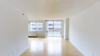 This single-floor space offers a clean, minimalist aesthetic defined by an open layout and a bright, airy atmosphere. The main living area is anchored by warm-toned light hardwood flooring that reflects natural light beautifully throughout the room, enhancing the sense of openness and space.

Large floor-to-ceiling sliding glass doors serve as a central architectural feature, inviting generous amounts of natural light and providing direct access to an outdoor balcony area. Flanking the sliding doors is an additional window, further maximizing the flow of daylight into the interior. The views look out onto surrounding urban architecture, giving the space a distinctly city-oriented character.

The walls are finished in a crisp, neutral white throughout, complementing the light palette of the floors and contributing to the clean, uncluttered feel of the space. A textured ceiling adds subtle dimension overhead. A built-in under-window heating unit is neatly integrated beneath the windows, maintaining the streamlined aesthetic of the room.

The open-plan layout provides excellent flexibility for a variety of living and furnishing arrangements. Overall, the space communicates a bright, modern, and functional character, well-suited for those seeking a light-filled urban environment with a straightforward, polished design sensibility.