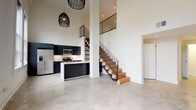This striking two-story space masterfully blends contemporary design with warm, organic materials. The main level immediately impresses with its soaring double-height ceilings that amplify the open, airy atmosphere. Polished concrete floors extend throughout, lending an industrial-chic sensibility to the living and kitchen areas. The kitchen is outfitted with bold dark cabinetry, stainless steel appliances including a side-by-side refrigerator, a gas range, and a built-in microwave, all anchored by a clean white island with an integrated sink. Statement woven pendant lights add a artisanal touch overhead. A sculptural staircase with natural wood treads and sleek black metal railings connects the two floors, serving as a focal architectural element. The main level also features a hallway leading to additional rooms, including a bathroom finished with distinctive hexagonal white mosaic tile surrounding a soaking tub, a dark floating vanity, and contrasting dark tile floors. Upstairs, the aesthetic shifts warmly with wide-plank reclaimed hardwood flooring throughout. The upper level includes generously sized bedrooms filled with natural light from large windows offering neighborhood views, a spacious walk-in closet with built-in wire shelving, and a second well-appointed bathroom echoing the hexagonal tile detailing found below. Clean white walls throughout both floors provide a crisp, neutral backdrop that highlights the thoughtful material choices at every turn.
