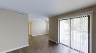 This single-floor residence showcases a cohesive, open layout that connects the living and adjoining areas seamlessly. The main living space is finished with rich, dark hardwood-style flooring that contrasts cleanly against the white baseboards and neutral taupe-toned walls, creating a warm yet understated aesthetic throughout. A ceiling fan is centrally positioned overhead, providing both functional airflow and a subtle design element to the room.

Natural light is a defining feature of the space, with large sliding glass doors fitted with vertical blinds along one wall. These doors open to an outdoor area, offering views of surrounding greenery and allowing ample daylight to fill the interior. The open sightlines from the main living area extend toward a hallway leading to additional rooms, suggesting a practical and well-organized floor plan. The transition between spaces is straightforward, with doorways visible in the background hinting at further rooms beyond. Overall, the space presents a clean, move-in-ready environment with durable finishes and a layout designed for comfortable everyday living.
