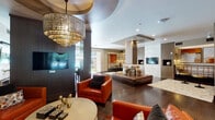 The Richdale Apartments clubhouse at 1901 S Voss Rd in Houston, Texas presents a thoughtfully designed, single-floor community space that blends contemporary sophistication with warm, inviting details. The glass-enclosed entry lobby sets an elegant tone, featuring polished tile floors, recessed lighting, and views of the lush outdoor pool courtyard lined with palm trees. Inside, rich hardwood floors flow throughout the main gathering areas, anchored by structural columns clad in warm wood and marble finishes. The central lounge is a standout, furnished with bold orange leather armchairs, leather ottomans, and plush textured seating arranged around a statement tiered chandelier with an ornate, layered design. A marble-surround wall niche houses a mounted flat-screen television, while curated abstract artwork adds vibrant color accents throughout. An intimate seating alcove with a coffered ceiling and a second dramatic chandelier provides a more private lounge setting. The kitchen area is impressively appointed, featuring an expansive marble-topped island with seating, warm wood cabinetry, beverage refrigerators, and a linear crystal pendant light overhead. White subway tile backsplash, a wall-mounted television, and gold-toned fixtures complete the space. The overall ambiance is upscale and modern, offering residents a refined environment for socializing and relaxation.