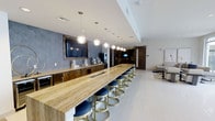 This single-floor space showcases a thoughtfully designed interior that blends contemporary style with functional versatility. The centerpiece is an expansive stone-topped bar counter finished in warm travertine tones, lined with a generous row of modern bar stools featuring gold-toned metal bases and navy blue upholstered seats. A series of geometric globe pendant lights hang above the bar, providing focused illumination and adding a decorative focal point to the space.

Along the back wall, rich dark wood cabinetry provides ample storage and counter space, complemented by a large wall-mounted television framed against a textured blue-gray accent wall. Built-in appliances are integrated seamlessly into the cabinetry, suggesting the space is equipped for entertaining or event hosting.

A sliding barn-style door in dark wood leads to an adjacent area, hinting at additional rooms beyond. The main space extends into a comfortable lounge area with seating, visible in the background. Light-toned polished flooring runs throughout, enhancing the bright, open atmosphere created by the white ceiling with recessed lighting. The overall ambiance is sleek, modern, and well-suited for social gatherings.
