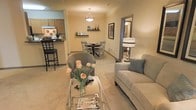 This single-floor residence showcases an open-concept design that integrates the living, dining, and kitchen areas into one cohesive and functional space. The living area is anchored by a contemporary sofa and a sleek glass coffee table, accented with fresh floral arrangements that add a welcoming touch. Neutral-toned flooring flows throughout, creating a sense of continuity across the space.

Adjacent to the living area, the dining space features a round table with X-back chairs positioned beneath a pendant light fixture, which adds a focal point to the room. Floating wall shelves display decorative accents, contributing to the curated aesthetic. A large mirror leaning against the wall enhances the sense of depth and light within the space.

The kitchen, visible from the main living area, includes a breakfast bar with bar seating, wood-toned cabinetry, and a tile backsplash, offering both functionality and style. A hallway leading from the living area hints at additional private rooms beyond. Overall, the space maintains a warm, neutral palette with modern furnishings, creating a comfortable and practical living environment.