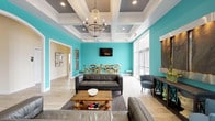 This single-story community space in Vineland makes a striking first impression with its bold teal accent walls, light wood-style flooring, and elegant coffered ceilings finished in white and gray. The entryway welcomes visitors with large glass double doors, a decorative wall-mounted water feature, and a console table with curated décor, setting a polished yet inviting tone.

The main lounge area is generously proportioned, anchored by a statement crystal chandelier suspended from the coffered ceiling. Dark leather sofas arranged around a rustic wood coffee table on a leopard-print area rug create a comfortable seating arrangement. Large windows flood the space with natural light and offer views of the surrounding community.

Adjacent to the lounge, a casual dining area features round bistro-style tables with cross-back bar stools and wall-mounted flat-screen televisions, making it suitable for both socializing and entertainment. Bold abstract artwork decorates the teal walls, adding visual interest.

The kitchen is well-appointed with gray shaker-style cabinetry, white countertops, stainless steel appliances, and a multi-seat island with industrial-style bar stools. The coffered ceiling continues here, maintaining design consistency throughout. Overall, the space balances energetic color with refined architectural detail.
