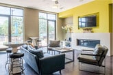 Located at 2764–2776 University Ave in Shorewood Hills, WI, this community lounge and kitchen is a well-appointed common area designed for comfort and social interaction. The open living area is anchored by rich hardwood flooring that runs throughout the space, complementing a warm yet contemporary color palette of taupe, chartreuse yellow, and crisp white accents. A sleek, linear fireplace serves as a focal point beneath a wall-mounted television set within a bold yellow accent niche, adding a modern touch to the gathering area. Abundant natural light pours in through dramatic floor-to-ceiling windows and glass doors, which open directly onto an outdoor terrace with patio seating — offering seamless indoor-outdoor connectivity. The lounge is furnished with leather seating, bar-height tables, and stools, while a foosball table and curated abstract artwork lend an energetic, social atmosphere. A ceiling fan enhances comfort in the main seating area. The adjacent kitchen is thoughtfully designed with crisp white cabinetry, a teal tile backsplash, light countertops, stainless steel appliances including a full-size refrigerator and built-in microwave, and a coffee station — making it fully equipped for communal use. Together, these spaces offer a functional and inviting environment ideal for residents to gather, relax, and connect.