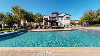 Located at 23820 Commercial Dr in Rosenberg, Texas, this single-story amenity space is designed to deliver an upscale, resort-inspired outdoor experience. The centerpiece is a generously sized swimming pool with crystal-clear blue water and a paver-lined deck, providing ample room for lounging and recreation under the Texas sun. Surrounding the pool, rows of chaise lounges offer comfortable sunbathing areas, while shade umbrellas provide relief from the heat.

A standout feature is the covered pavilion, constructed with striking natural stone columns and a warm wood-paneled ceiling with ceiling fans. The pavilion hosts a substantial dining table with wrought iron and cushioned seating, making it well-suited for outdoor gatherings. Large glass-paned double doors connect the pavilion to the interior, while open sightlines to the pool and landscaping enhance the sense of openness.

An additional pergola structure offers a shaded retreat with modern lounge seating and views of palm trees and manicured grounds. The exterior architecture blends light-toned limestone masonry with dark-framed windows and doors, creating a refined, cohesive aesthetic throughout the outdoor space.
