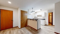 This single-floor unit offers a clean, functional layout designed for comfortable everyday living. The open living area is finished with durable wood-style flooring that runs throughout the main living spaces, complemented by neutral white walls and baseboard heating. A large window at the far end of the living room fills the space with natural light and frames views of surrounding greenery, creating a bright and welcoming atmosphere.

The kitchen is efficiently designed with white cabinetry accented by sleek black hardware, dark countertops, and light wood-style flooring. A peninsula with an overhang provides casual seating and separates the kitchen from the living area, while pendant lighting adds a modern touch overhead. Full-size appliances, including a refrigerator, stove, and dishwasher, are included, along with ample upper and lower cabinet storage and a double basin sink.

The bedroom is a calm, carpet-floored retreat featuring a window with a view of mature trees, offering a quiet and private feel. Baseboard heating keeps the room comfortable year-round.

The bathroom is finished in clean, all-white tones with a combined tub and shower unit, a curtain rod, and chrome fixtures, maintaining a simple and practical aesthetic throughout. Warm-toned interior doors add a natural wood element that ties the unit's design together cohesively.