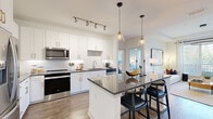 This well-appointed single-floor residence showcases a thoughtfully designed open-concept layout that seamlessly connects the kitchen, dining, and living areas. The kitchen serves as a central highlight, featuring floor-to-ceiling white shaker cabinetry with brushed gold and silver hardware, dark granite countertops, a textured tile backsplash, and a full suite of stainless steel appliances. A substantial kitchen island with seating for multiple guests provides additional prep space and a natural gathering point. Pendant lighting and track fixtures illuminate the space with warm, layered light.

The living area benefits from a tray ceiling with a modern circular light fixture, wood-look flooring, and large sliding glass doors and windows that draw in abundant natural light and provide views of the surrounding greenery and exterior balcony access.

Two well-proportioned bedrooms feature plush carpeting, large windows with blinds, and a calm, neutral palette. The primary bathroom offers a dual-sink vanity with dark granite countertops, white cabinetry, a large mirror, and an adjoining shower with marble-style tile. A secondary bathroom mirrors the same refined finishes with a single vanity and granite countertop, maintaining a consistent, polished aesthetic throughout the residence.