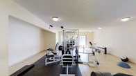 This fitness center offers a comprehensive workout environment designed to support a variety of exercise routines. The space is laid out on a single floor with a clean, functional aesthetic defined by light-colored walls and tile flooring accented by rubber mat sections beneath the equipment for safety and stability.

At the heart of the room stands a multi-station cable machine, providing a wide range of resistance training options. Surrounding it is an assortment of strength and conditioning equipment, including leg press and extension machines, weight benches, and free weights, making the facility suitable for both targeted muscle training and general fitness.

Natural light filters into the space through a large window at the far end, contributing to an open and energizing atmosphere. Overhead flush-mount lighting ensures the space remains well-lit throughout. The textured ceiling adds a modest architectural detail that complements the no-frills, purpose-driven design of the room.

The layout is thoughtfully organized, providing clear circulation paths between pieces of equipment while maximizing the available floor space. Overall, this fitness center delivers a practical and inviting environment well-suited for residents or guests seeking a convenient on-site workout facility.