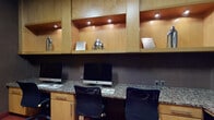This single-floor business center is thoughtfully designed to support productivity and professional needs. The main workstation area features a continuous granite-topped desk with multiple Apple iMac computers, ergonomic mesh office chairs, and built-in wood cabinetry above, complete with recessed accent lighting and open display shelves adorned with decorative metallic accents. The warm maple wood tones of the cabinetry contrast elegantly with the dark textured wall coverings, creating a focused yet polished atmosphere. A dedicated private workstation alcove offers additional workspace, complete with its own computer, a multifunction printer/copier, and under-counter storage — ideal for more confidential or extended work sessions. This alcove is enclosed by a glass-paneled wood door, providing privacy while maintaining visual connection to the broader space. A comfortable seating area with wood-framed upholstered chairs is positioned near large interior windows that look out to an adjacent corridor with marble-tiled walls and elegant ceiling fixtures, adding a touch of refinement to the overall environment. Red carpeting throughout ties the space together, complementing the warm wood and dark wall tones. Framed abstract artwork adds a creative touch, rounding out a space that is both functional and visually inviting.