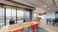 Located at 3029 22nd Ave S in Minneapolis, MN, this rooftop amenity space offers a dynamic blend of indoor comfort and outdoor living. The interior is defined by eye-catching stamped tin ceilings, exposed industrial ductwork, and a mix of herringbone wood and textured tile flooring that creates a stylish, urban aesthetic. A spacious lounge area features floor-to-ceiling glass walls that frame panoramic views of the Minneapolis skyline, filled with vibrant orange seating, colorful rugs, and abundant natural light. Adjacent to the lounge, a communal dining area centers around a large wood table with bold red chairs, integrated planter boxes, and direct sliding glass door access to the terrace. The game and recreation zone includes a foosball table and vending machines, making it a lively gathering space. A well-appointed kitchenette offers granite countertops, warm wood cabinetry, a double sink, dishwasher, and a wall-mounted television. The outdoor terrace is a standout feature, equipped with a pergola structure, a long linear fire pit, raised planter beds with lush greenery, and Adirondack-style seating—all set against sweeping rooftop views of the surrounding Minneapolis neighborhood.