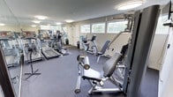 This fitness center offers a comprehensive workout environment designed to support a wide range of exercise routines. The space is finished with durable dark carpet flooring and benefits from natural light streaming in through a row of horizontal windows that line the upper portion of the walls, providing views of the surrounding neighborhood and greenery. Flush-mount ceiling lights supplement the natural light, ensuring the space remains well-lit throughout the day. A full-length wall mirror expands the visual depth of the room while allowing users to monitor their form during workouts. The cardio section is prominently featured, housing multiple treadmills and elliptical trainers, as well as upright and recumbent stationary bikes suited for varying fitness levels. A dedicated strength training area includes a multi-station cable machine, ideal for a variety of resistance exercises, along with a flat weight bench and a well-stocked dumbbell rack. A standing fan provides additional ventilation and comfort during intense workouts. A wall-mounted television adds an entertainment option for users. A water cooler is conveniently positioned near the entrance, and a private door leads to an adjacent room. The overall layout is organized and practical, making the fitness center a fully functional facility for both cardio and strength training needs.