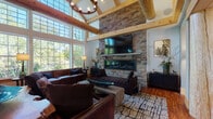This single-floor space showcases an impressive blend of rustic craftsmanship and modern comfort. The expansive great room commands attention with its soaring vaulted ceiling, exposed timber beams, and a dramatic floor-to-ceiling stacked stone fireplace topped by a mounted flat-screen TV. Large multi-pane windows flood the space with natural light and offer sweeping views of the surrounding tree canopy, while rich hardwood flooring runs throughout. Deep leather seating and patterned area rugs create a warm, inviting atmosphere anchored by the fireplace.

The kitchen is a standout feature, designed with bold navy cabinetry, granite countertops, stainless steel appliances, and a functional center island — all accented by exposed timber beams overhead. A welcoming entry area features a live-edge wood table with resin accents, artisan seating, and warm pendant lighting.

A dedicated fitness room is bathed in natural light from an array of large windows and is equipped with treadmills, an elliptical, free weights, and floor mirrors — ideal for a full workout routine. A serene lounge or relaxation room with soft neutral tones, decorative mirrors, and plush seating completes this thoughtfully appointed space, offering a retreat for rest and recovery.